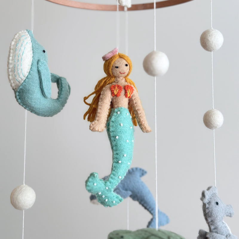 A felt baby mobile, featuring a mermaid and various sea creatures, hangs from a string, suspended in mid-air. The mobile is made of wool felt and has a serene, tranquil atmosphere, evoking the beauty of marine life. The product's neutral background allows the mobile to be the focal point, suggesting it would be used to decorate a nursery or child's room.