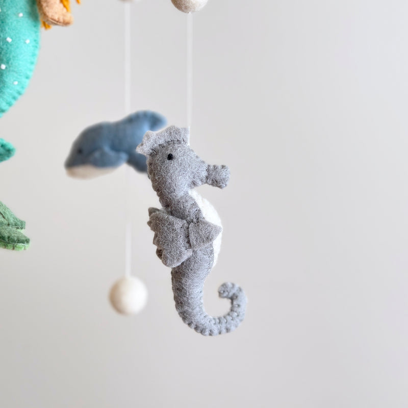 A handmade wool felt baby mobile, featuring a mermaid and various sea creatures, hangs against a light background. The soft pastel colors bring an underwater world to life. Made from 100% wool felt, this unique mobile is suspended from a string, creating a serene ambiance for your nursery.