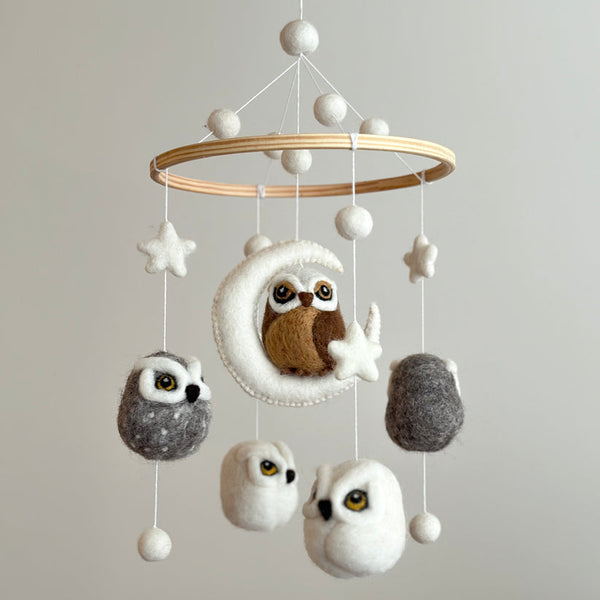 A whimsical wool felt baby mobile, featuring a wooden frame supporting a collection of colorful felt owls in various sizes, suspended from the ceiling by a string. The central brown owl has large eyes, while smaller unique white and gray owls surround it, creating a warm and playful atmosphere.