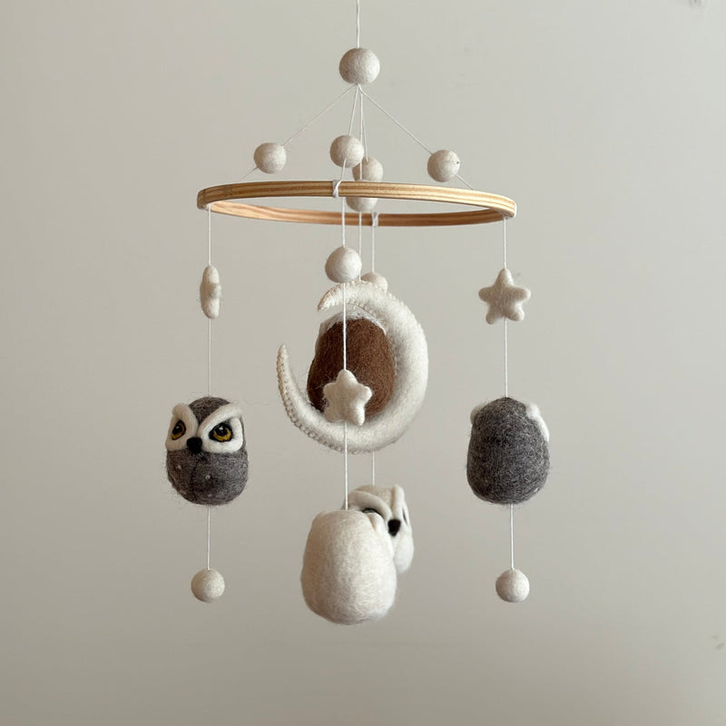 A charming wool felt baby mobile, featuring an owl as the central figure, resting on a wooden table. The mobile's wooden frame adds stability and durability, while its whimsical design includes a moon, stars, and small creatures surrounding the owl. The background is clean and minimalist, showcasing the vibrant details of the mobile.