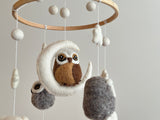 A charming wool felt owl baby mobile hangs from a wooden frame, suspended above a white wall. The mobile features an owl and several fish-shaped felt sea creatures, each attached to cotton thread. The product is made of wool felt and has a playful, whimsical design.