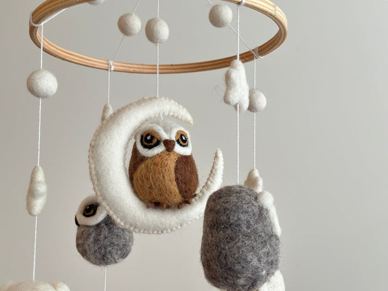 A charming wool felt owl baby mobile hangs from a wooden frame, suspended above a white wall. The mobile features an owl and several fish-shaped felt sea creatures, each attached to cotton thread. The product is made of wool felt and has a playful, whimsical design.