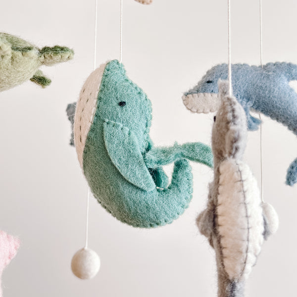 A close-up of a wool felt Sea Life baby mobile features several suspended sea creatures against a plain white background, creating a serene atmosphere. A light-green whale with a white belly hangs centrally, with a gray sea horse to its right. Above, a blue dolphin and a partially visible light-green sea turtle complete the tranquil ocean theme.