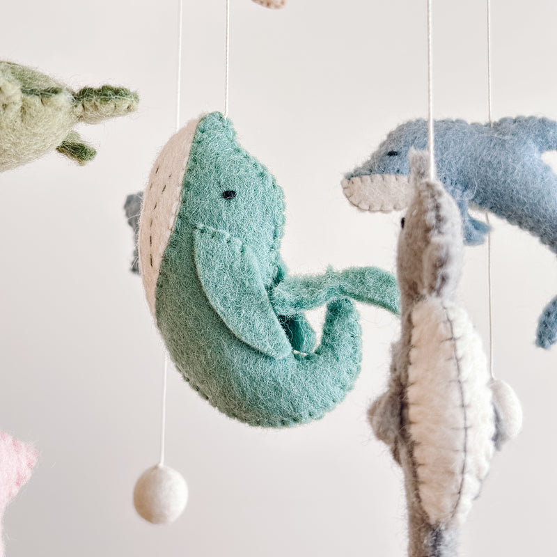 A close-up of a wool felt Sea Life baby mobile features several suspended sea creatures against a plain white background, creating a serene atmosphere. A light-green whale with a white belly hangs centrally, with a gray sea horse to its right. Above, a blue dolphin and a partially visible light-green sea turtle complete the tranquil ocean theme.
