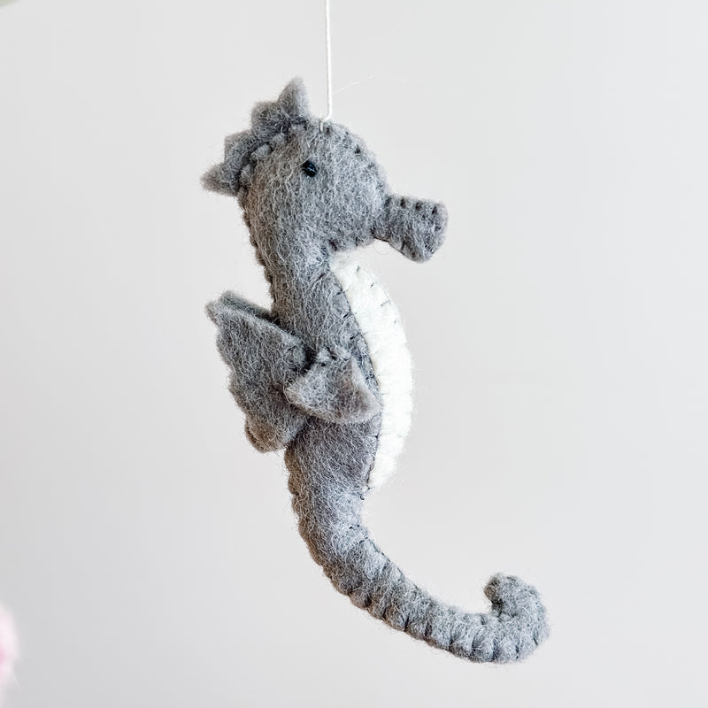 A gray wool felt seahorse, part of a sea life baby mobile, hangs centrally from a thin white string within a wooden frame against a plain white background. The seahorse features a distinctive curled tail and a white belly, with a visibly soft, plush felt texture.