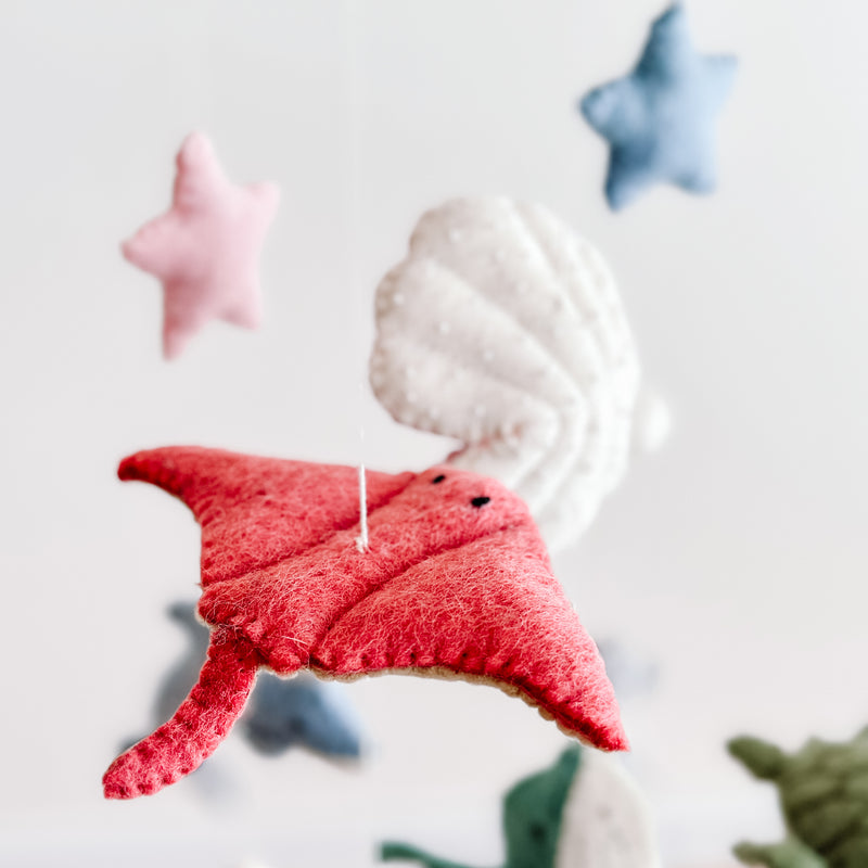 A red wool felt stingray, part of a whimsical sea life baby mobile, is prominently featured against a soft, off-white background. Crafted from textured red felt, the stingray is suspended by a thin white cotton thread and features small black button eyes. A visible wooden frame and partially seen wool felt scallop complete the playful, serene oceanic scene.