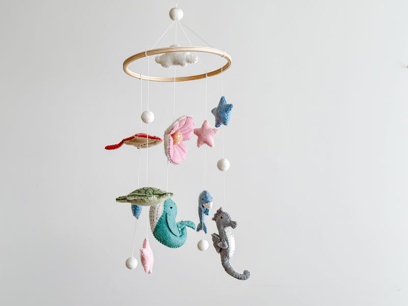 A Sea Life wool felt baby mobile is displayed against a plain white background, creating a calm atmosphere. It features a wooden ring from which a green sea turtle, a teal whale, a blue dolphin, and a grey seahorse are suspended by thin white cotton threads. White wool felt balls are positioned at the top, in the center of the ring, and at the bottom of some threads, adding a playful touch perfect for a nursery.
