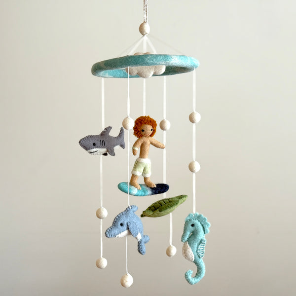 A Surfer Boy & Ocean Creatures felt baby mobile is displayed against a light gray background, creating a calm, playful atmosphere. The handcrafted mobile features felt figures of a surfer boy with orange hair on a blue and black surfboard, a green sea turtle, a blue dolphin, and a light blue seahorse. These hang from a round, light blue felt ring by white strings, interspersed with felt balls. A braided white cord with a felt ball provides easy hanging.