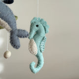 A wool felt baby mobile featuring ocean creatures, a seahorse, whale, and a round felt ball, is displayed against a plain off-white background. The central light blue felt seahorse has a cream belly patch and delicate white stitching for its gills. A blue and cream whale is partially visible alongside a round felt ball, all crafted from soft felted wool, lending a handmade aquatic theme.