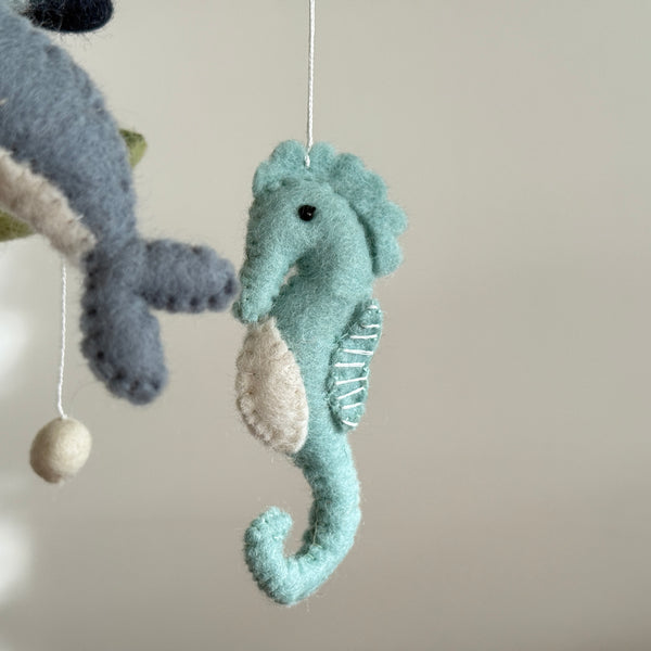A wool felt baby mobile featuring ocean creatures, a seahorse, whale, and a round felt ball, is displayed against a plain off-white background. The central light blue felt seahorse has a cream belly patch and delicate white stitching for its gills. A blue and cream whale is partially visible alongside a round felt ball, all crafted from soft felted wool, lending a handmade aquatic theme.