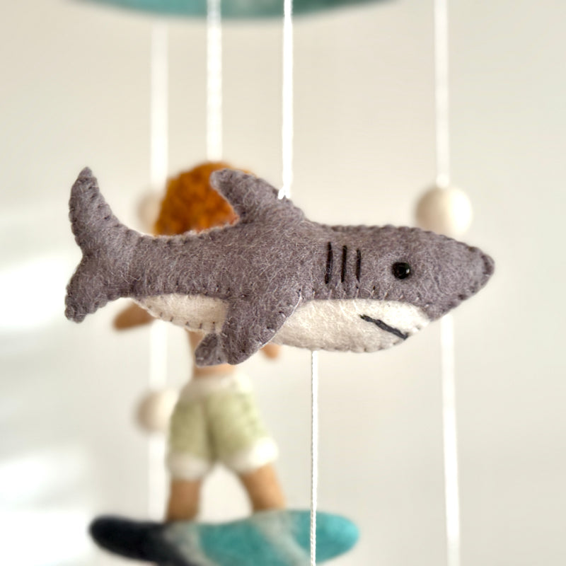 A whimsical wool felt baby mobile from the Surfer Boy & Ocean Creatures collection is suspended in a close-up studio shot against a plain white backdrop. It features a playful felt surfer boy with orange hair standing on a vibrant teal surfboard. White felt balls adorn the mobile's strings, enhancing its decorative appeal.