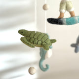 A light-green felt sea turtle, part of the Surfer Boy & Ocean Creatures baby mobile, is captured in a close-up against a soft, neutral backdrop. This plush felt ocean creature features detailed stitching that mimics a natural turtle shell, creating a serene, floating appearance. Softly blurred elements like a surfer boy and seahorse from the mobile are visible in the background.