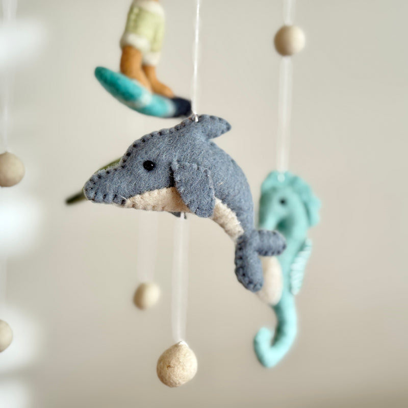 A delightful Surfer Boy and Ocean Creatures felt baby mobile hangs in a close-up studio shot against a soft beige background. This serene mobile showcases a gray felt dolphin with a cream underbelly and stitched features, a light turquoise felt seahorse with a curved tail, and a surfer riding a turquoise board. All components are suspended by white strings accented with light beige wooden beads, creating a whimsical ocean scene.