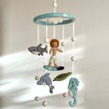 A Surfer Boy and Ocean Creatures felt baby mobile hangs in the center of a softly lit room against a neutral background. It features a circular, light blue felt frame with a single felt ball above it. Handcrafted felt figures are suspended below: a surfer boy with orange hair on a blue and black surfboard, a gray dolphin, a dark green sea turtle, and a light blue seahorse. Small felt balls are placed between the figures.