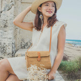 A woman stands outdoors against a stone wall and a body of water, wearing a light-colored dress and straw hat. She holds a large rattan crossbody bag with a leather strap, fitting comfortably on her shoulder. The Bali Messenger Rattan Crossbody Bag - Large is prominently displayed, showcasing its natural color and texture blending well with the outdoor setting.
