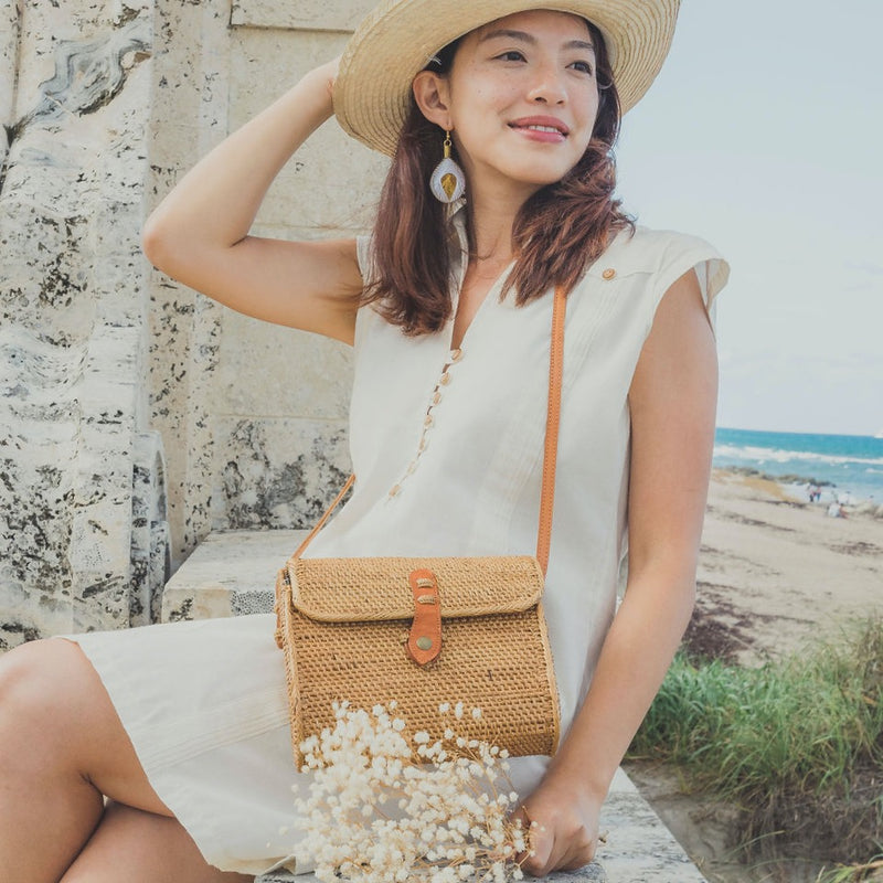 A woman stands outdoors against a stone wall and a body of water, wearing a light-colored dress and straw hat. She holds a large rattan crossbody bag with a leather strap, fitting comfortably on her shoulder. The Bali Messenger Rattan Crossbody Bag - Large is prominently displayed, showcasing its natural color and texture blending well with the outdoor setting.