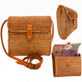 A woven rattan crossbody bag rests on a wooden surface, showcasing its rectangular shape and floral pattern. The bag features adjustable brown leather straps and two pockets secured by button closures, giving it a rustic yet stylish appearance.
