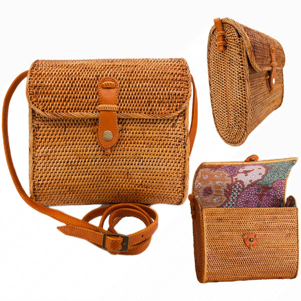 A woven rattan crossbody bag rests on a wooden surface, showcasing its rectangular shape and floral pattern. The bag features adjustable brown leather straps and two pockets secured by button closures, giving it a rustic yet stylish appearance.