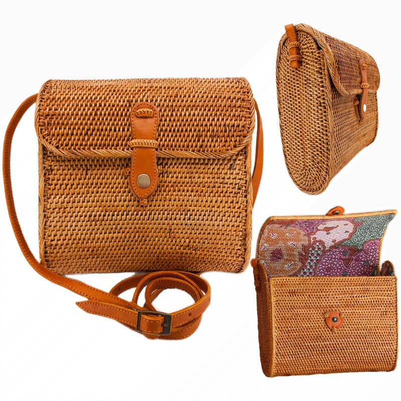 A woven rattan crossbody bag rests on a wooden surface, showcasing its rectangular shape and floral pattern. The bag features adjustable brown leather straps and two pockets secured by button closures, giving it a rustic yet stylish appearance.