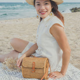 A woman sits on the sand at a beach, wearing a white blouse and straw hat. In front of her is a woven bag with a brown handle, crafted from Ata grass with a unique Batik lining.