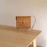 A small, woven rattan purse rests on a wooden table, showcasing its light brown color and simple rectangular shape. The flap closure is secured by a metal buckle, and the background features a plain wall and white door frame, suggesting an indoor setting.