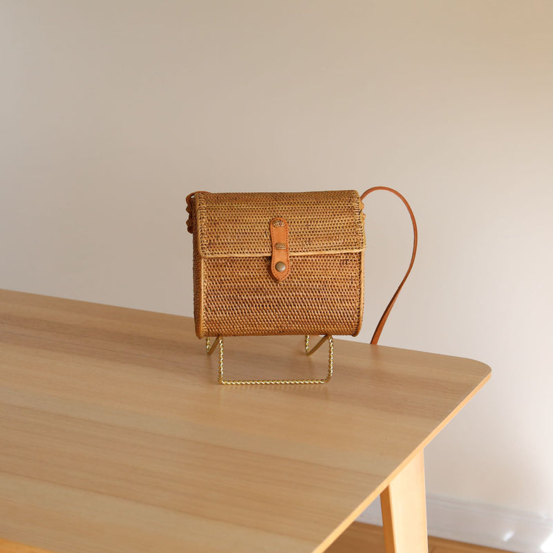 A small, woven rattan purse rests on a wooden table, showcasing its light brown color and simple rectangular shape. The flap closure is secured by a metal buckle, and the background features a plain wall and white door frame, suggesting an indoor setting.