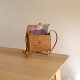 A small, woven rattan purse rests on a wooden surface, showcasing its textured appearance and colorful pattern featuring shades of blue, green, and pink. The purse has a flap closure and a small rectangular tag attached to the front, suggesting it's made from natural materials like rattan or Ata grass. This casual, relaxed image focuses on the purse as the main subject.