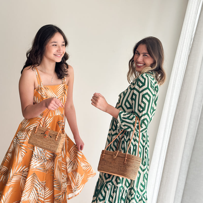 A woman wearing a bright yellow dress stands on the left, smiling at the camera, while another woman on the right holds a large woven bag made of rattan with a leather handle. The bag features a smooth finish and is lined with Batik cotton, with a snap closure. The background is minimalistic, focusing attention on the women and their accessories.