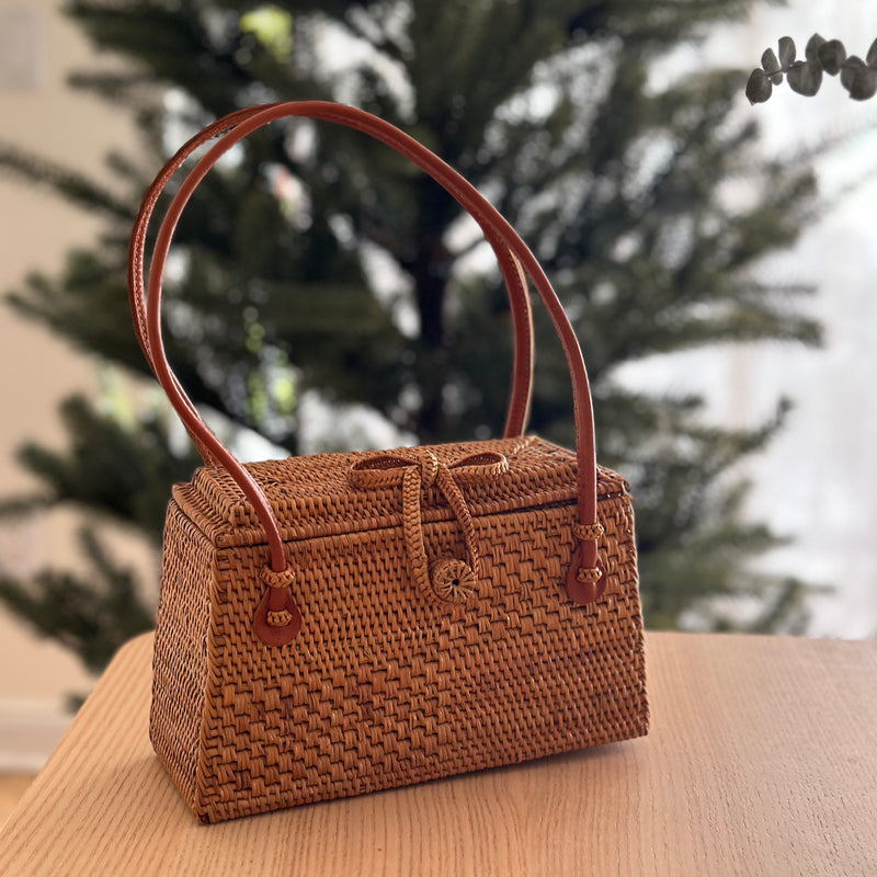A woven rattan handbag with a brown leather handle rests on a table, surrounded by a festive holiday setting. The trapezoid-shaped bag features a smooth, polished rattan finish, a unique Batik cotton lining, and a secure bowtie clasp. Made from sustainable materials, this eco-friendly and fair-trade product is handmade in Bali.