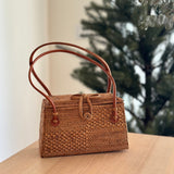A woven rattan bag, the Bali Rattan Bag - Maya, rests on a wooden surface. The brown-colored bag features a leather handle and bowtie clasp with secure snaps for closure. Set against a festive holiday background, the product showcases its smooth finish, secure closure, and leather handle.