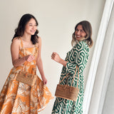 A cheerful, relaxed atmosphere is set by two individuals standing side by side against a plain background. They are each holding a Bali Maya Rattan Bag with a smooth finish, leather handle, and bowtie clasp. The bag's trapezoid shape and unique Batik cotton lining are visible, showcasing its elegant design.