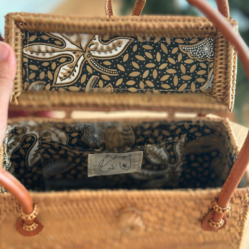 A person holds an open woven rattan bag with a floral batik lining pattern, made from natural materials like rattan. The bag features a bowtie clasp closure and a leather handle on the top edge, showcasing its design and craftsmanship. The setting is indoor, with a wall in the background.