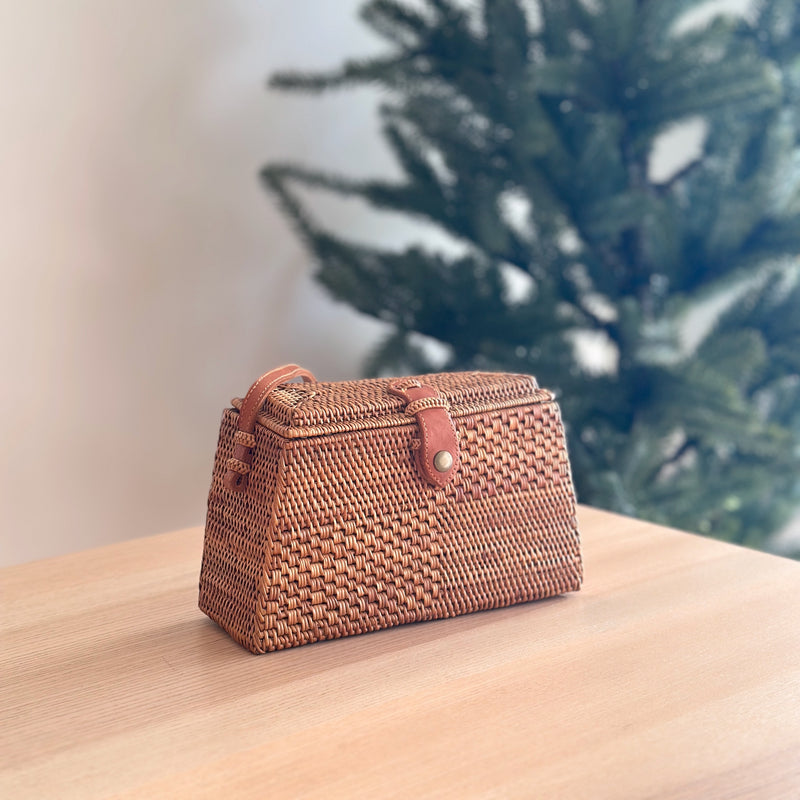 A Bali Rattan Crossbody Bag, titled 'Maya', rests on a wooden surface, showcasing its rattan material with a basket weave pattern. The bag features a rectangular shape with rounded corners, a flap closure secured by a leather snap button, and an adjustable strap for versatile wear. This stylish accessory complements the festive setting of a Christmas tree in the background.
