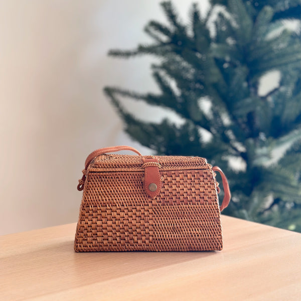 A woven rattan crossbody bag, Maya model, rests on a wooden table, its light brown color complementing the festive Christmas tree background with decorations. The bag features a centered leather snap clasp and an adjustable strap, showcasing its handcrafted quality with a textured surface made from natural materials like bamboo or rattan.