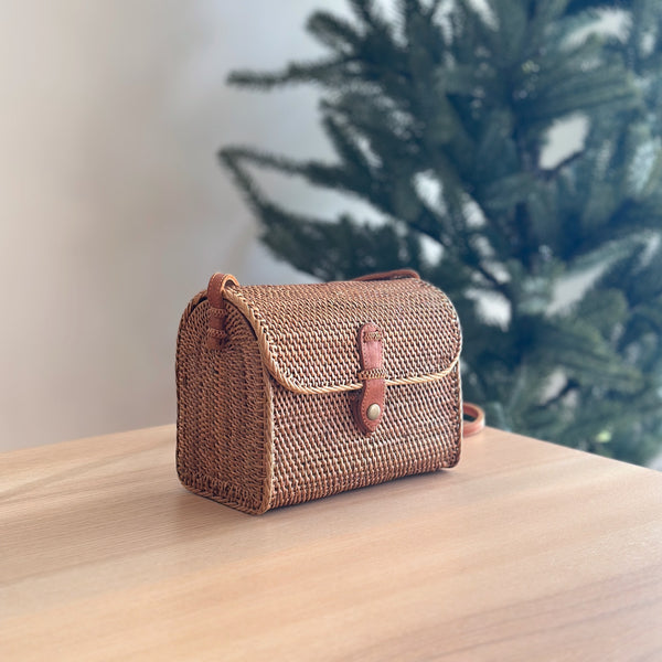 A woven rattan crossbody bag, Bali Rattan Crossbody Bag - Joey, rests on a wooden surface, evoking a warm and festive atmosphere. The bag's natural materials give it an organic feel, with a casual yet stylish design that exudes bohemian charm. Made from rattan and featuring a leather snap clasp, the bag boasts a snag-free finish and elegant silhouette. Perfect for effortless style, this eco-friendly accessory is handmade by Balinese artisans in Bali.