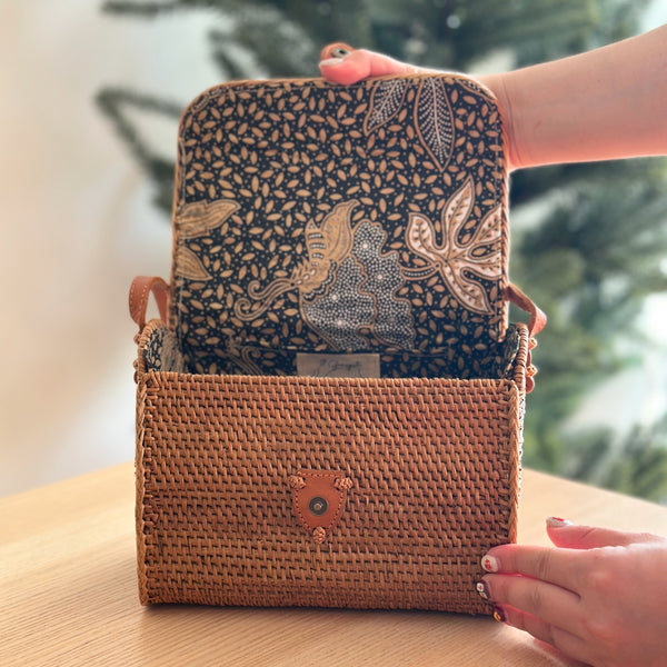 A person's hand holds an open Bali Rattan Crossbody Bag - Joey, showcasing its rattan material and floral pattern. The bag has a rectangular shape with rounded corners, featuring a central leather snap clasp and Batik cotton lining.