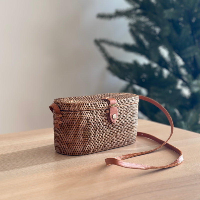 A woven crossbody bag crafted from Ata grass and real leather, resting on a surface in a cozy indoor setting. The rectangular shape features rounded corners, an adjustable brown leather strap for carrying over the shoulder, and a compact design. Made from environmentally friendly materials, this handcrafted piece exudes timeless elegance.