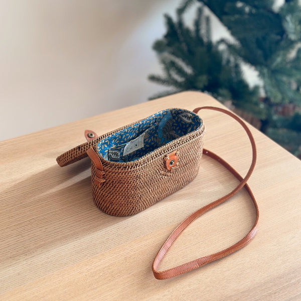 Bali Rattan Crossbody Bag - Sundung resting on a wooden surface, featuring a woven texture predominantly brown with blue accents. The bag has a decorative flower or emblem on the front, an adjustable strap made from real leather matching its color.