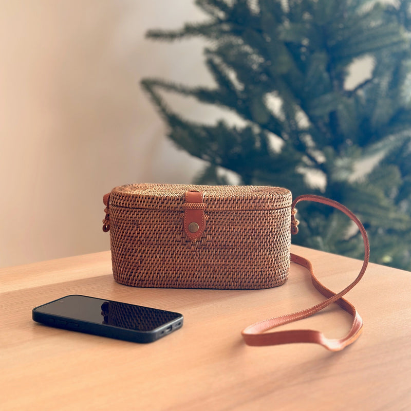 A woven crossbody bag, crafted from natural materials rattan or Ata grass, rests on a wooden surface. The textured finish features a small top handle and an adjustable strap that wraps around the body of the bag. The bag's natural texture and practical design evoke a cozy atmosphere, perfect for carrying personal items like a phone.