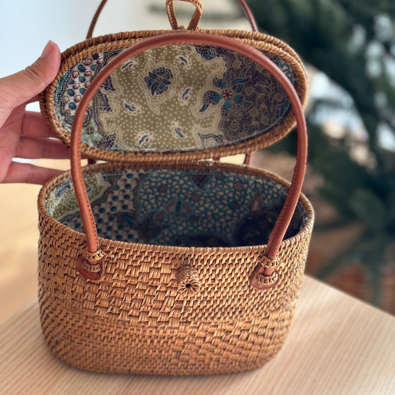 A Bali Rattan Handbag - Athens, resting on a table with its woven texture and predominantly brown and beige colors visible. The bag's interior is open, showcasing the unique Batik cotton lining in blue and white hues. A leather handle adorns the top, adding a touch of sophistication.