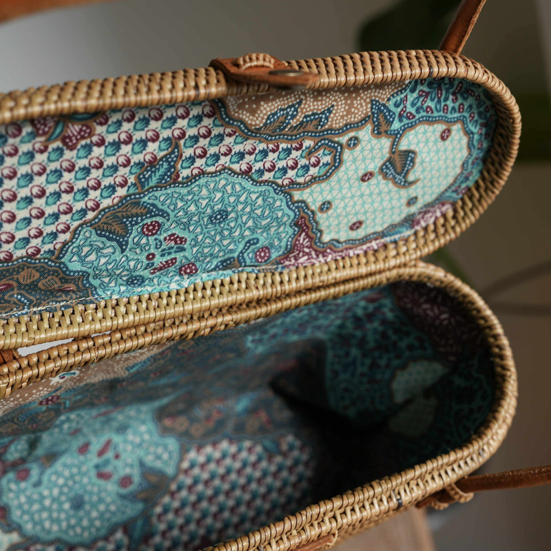 An inside look at a handwoven rattan shoulder bag reveals its exquisite, colorful Batik lining. The intricate, traditional pattern in shades of turquoise, blue, and brown adds a hidden layer of artistry to the natural Ata grass weave. The open lid showcases the detailed craftsmanship and the unique fabric that makes each bag a one-of-a-kind piece.