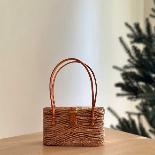 A Bali Rattan Handbag - Catania, resting on a surface in an indoor setting. The handbag is made of rattan with a woven texture and features brown handles attached to the top. It has a casual yet stylish design with a relaxed silhouette, suitable for various occasions.