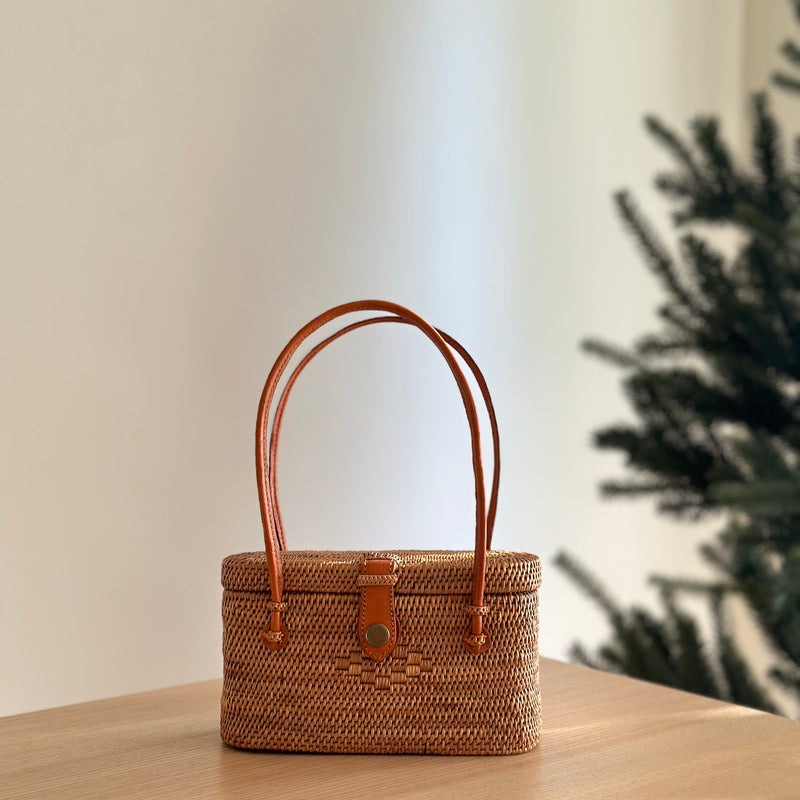 A Bali Rattan Handbag - Catania, resting on a surface in an indoor setting. The handbag is made of rattan with a woven texture and features brown handles attached to the top. It has a casual yet stylish design with a relaxed silhouette, suitable for various occasions.