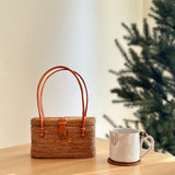 A woven rattan handbag, Bali Rattan Handbag - Catania, rests on a wooden surface next to a white mug and small Christmas tree. The bag features real leather handles, secure snap closure, and Batik cotton lining. Its smooth finish and snug fit showcase durability and comfort.