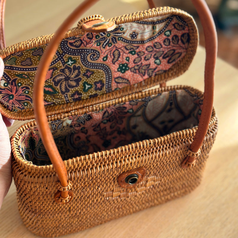 A petite rattan handbag, Catania, resting on a woven mat, showcasing its smooth finish and real leather handles. The bag features a unique Batik cotton lining and is made from eco-friendly materials.