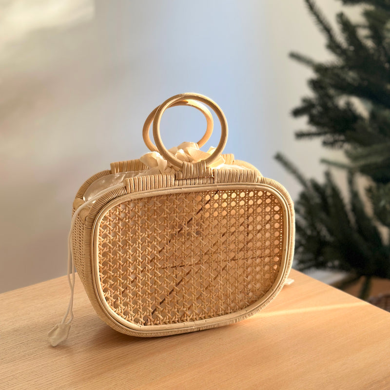 A woven handbag with a natural rattan handle sits on a wooden surface, surrounded by a Christmas tree backdrop and a section of wall. The bag's eco-friendly materials and unique design elements, such as batik lining and drawstring pouch, are highlighted. The overall mood is warm and inviting, suggesting a festive atmosphere.