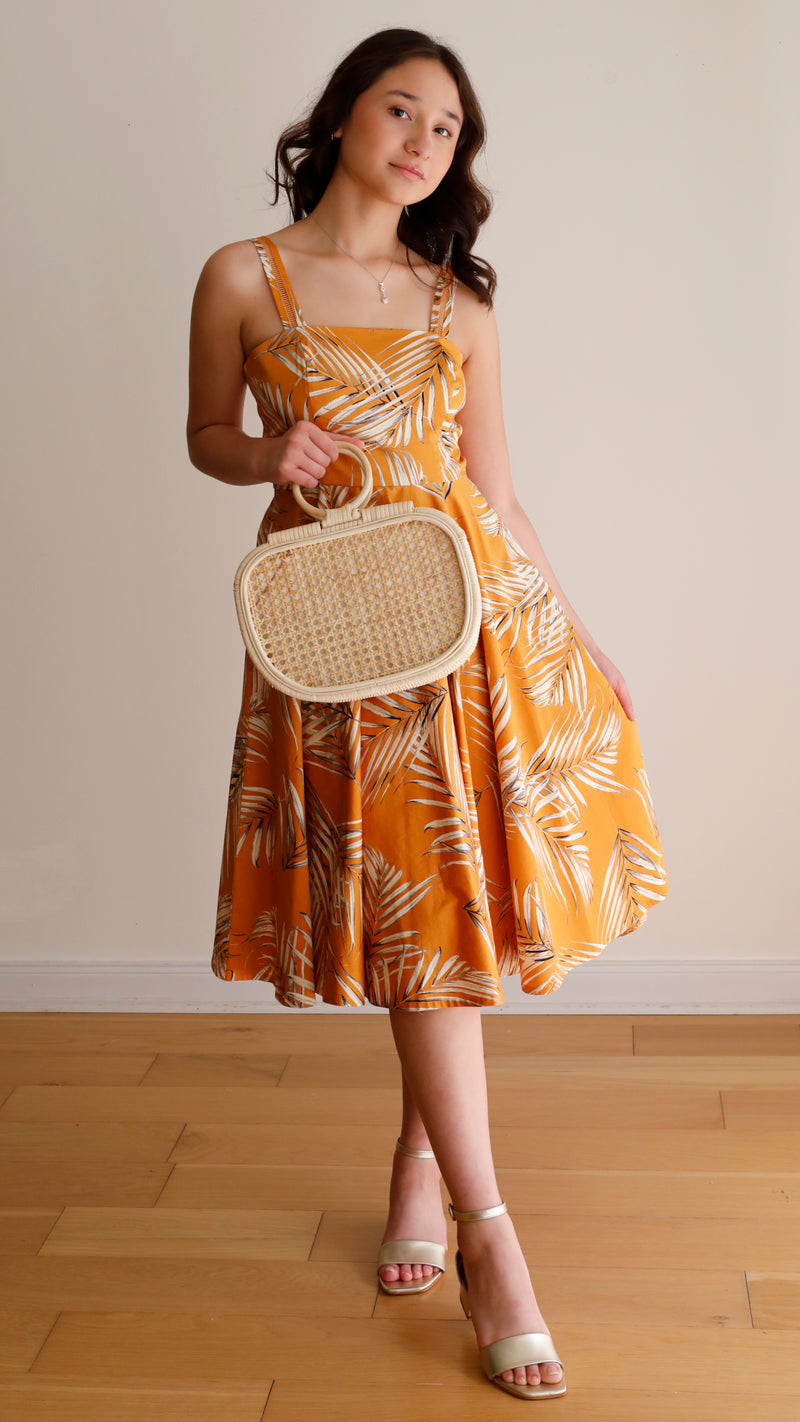 A woman stands against a plain background, wearing a bright orange dress with a floral pattern and styled hair. She holds a small, woven purse made from natural materials like rattan or similar, featuring a textured surface and drawstring closure.