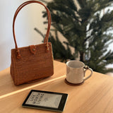 A woven rattan handbag with a leather snap clasp on top, placed on a wooden surface next to a coffee cup. The brown-colored handbag has a tulip-like silhouette when viewed from the side and appears to be made of smooth material with a polished finish.