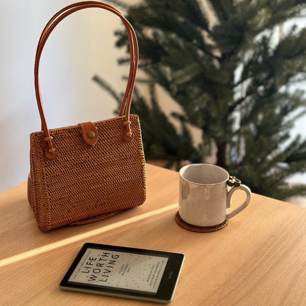 A woven rattan handbag with a leather snap clasp on top, placed on a wooden surface next to a coffee cup. The brown-colored handbag has a tulip-like silhouette when viewed from the side and appears to be made of smooth material with a polished finish.