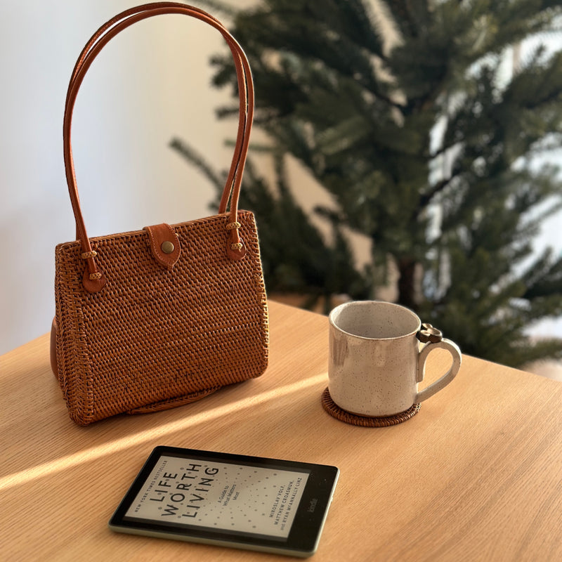 A woven rattan handbag with a leather snap clasp on top, placed on a wooden surface next to a coffee cup. The brown-colored handbag has a tulip-like silhouette when viewed from the side and appears to be made of smooth material with a polished finish.